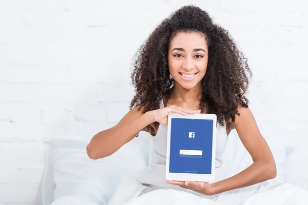 Glückliche afrikanisch-amerikanische Mädchen zeigt digitales Tablet mit Facebook auf dem Bildschirm im Bett zu Hause — Stockfoto