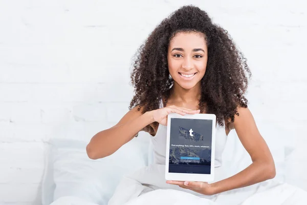 Menina americana africana feliz mostrando tablet digital com tumblr na tela na cama em casa — Fotografia de Stock