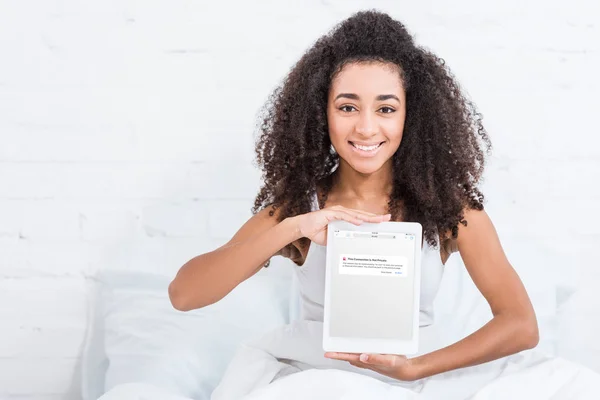 Happy african american girl showing digital tablet with blocked vk.com on screen in bed at home — Stock Photo