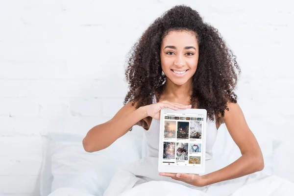 Happy african american girl showing digital tablet with pinterest on screen in bed at home — Stock Photo