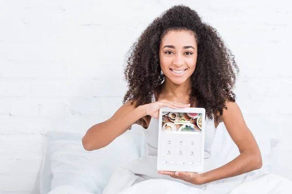 Chica afroamericana feliz mostrando tableta digital con cuadrado en la pantalla en la cama en casa - foto de stock