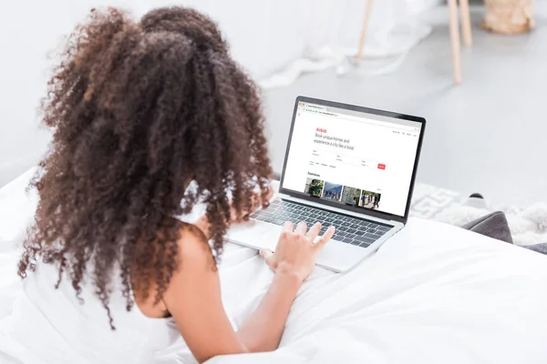 Rear view of curly woman using laptop with airbnb on screen in bed at home — Stock Photo