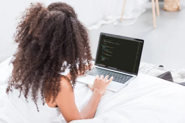 Vue arrière de la femme bouclée à l'aide d'un ordinateur portable avec code de programmation à l'écran dans le lit à la maison — Photo de stock