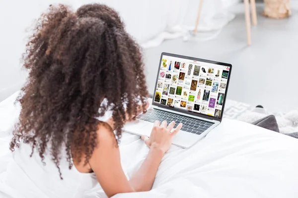 Vue arrière de la femme bouclée en utilisant un ordinateur portable avec pinterest à l'écran dans le lit à la maison — Photo de stock