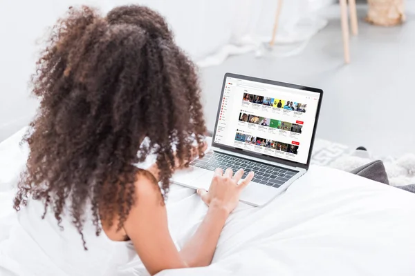 Rear view of curly woman using laptop with youtube on screen in bed at home — Stock Photo