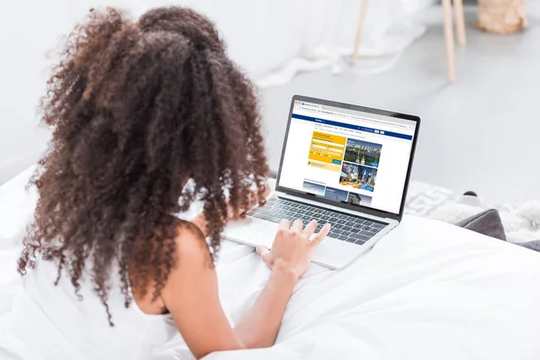 Rear view of curly woman using laptop with booking on screen in bed at home — Stock Photo