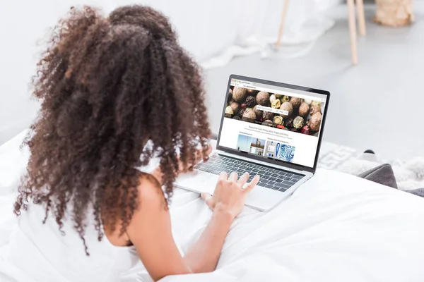 Rückansicht der lockigen Frau mit Laptop mit depositphotos.com auf dem Bildschirm im Bett zu Hause — Stockfoto