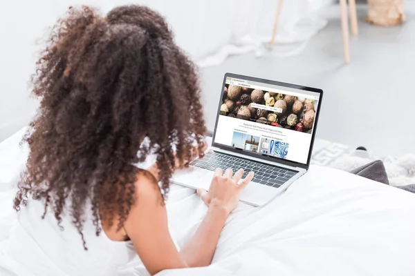 Vue arrière de la femme bouclée à l'aide d'un ordinateur portable avec depositphotos.com à l'écran au lit à la maison — Photo de stock