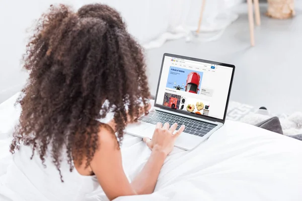 Rear view of curly woman using laptop with ebay on screen in bed at home — Stock Photo