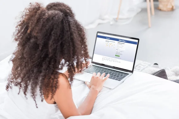 Rückansicht einer lockigen Frau mit Laptop und Facebook auf dem Bildschirm im heimischen Bett — Stockfoto
