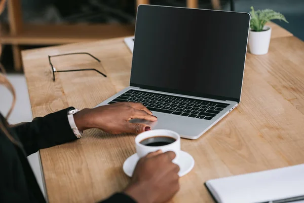 Cropped view of african american woman holding cup of coffee near laptop with blank screen — Stock Photo