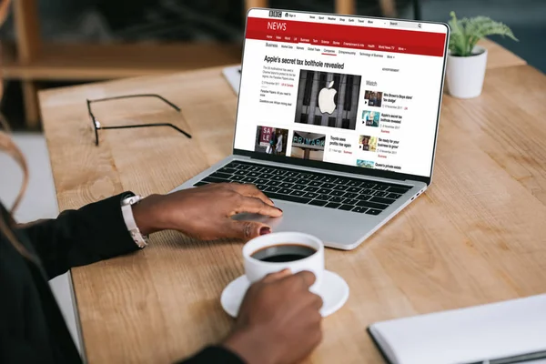 Vista ritagliata della donna afro-americana utilizzando il computer portatile con sito web di notizie vicino a tazza di caffè — Foto stock