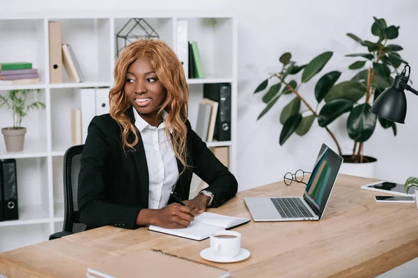 Selektiver Fokus einer fröhlichen und nachdenklichen afrikanisch-amerikanischen Geschäftsfrau, die in Notizbuch schreibt — Stockfoto