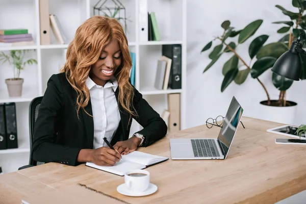 Glückliche afrikanisch-amerikanische Geschäftsfrau schreibt in Notizbuch — Stockfoto