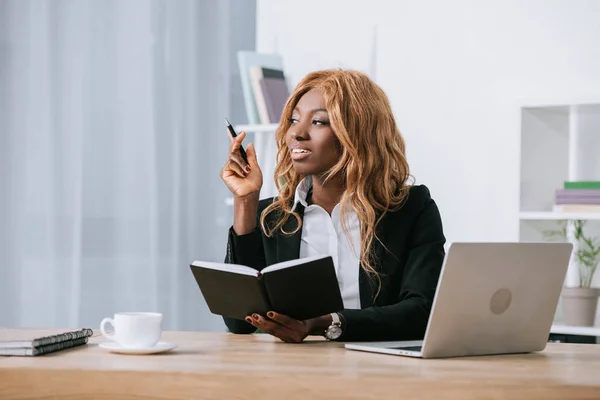 Bela mulher de negócios afro-americana segurando notebook e caneta no escritório moderno — Fotografia de Stock