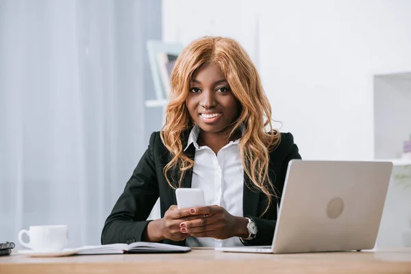 Африканская американская бизнесвумен держит смартфон рядом с ноутбуком в офисе — стоковое фото