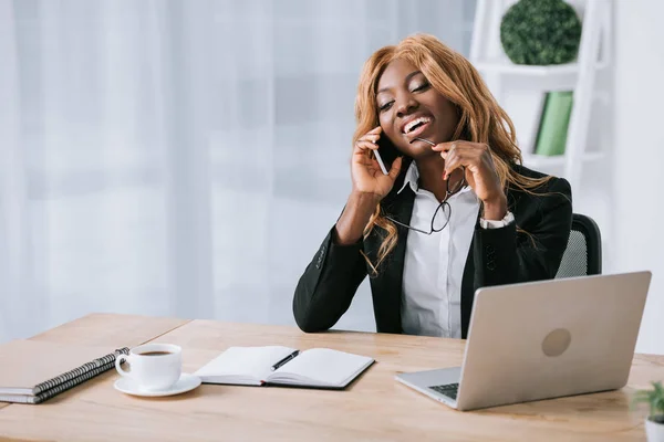 Alegre mujer de negocios afroamericana hablando en el teléfono inteligente en la oficina - foto de stock