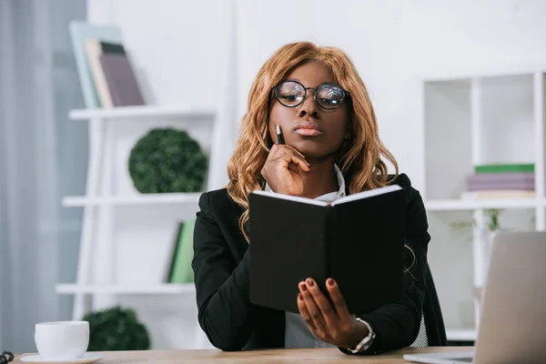 Pensativa mujer de negocios afroamericana sosteniendo cuaderno y pluma - foto de stock
