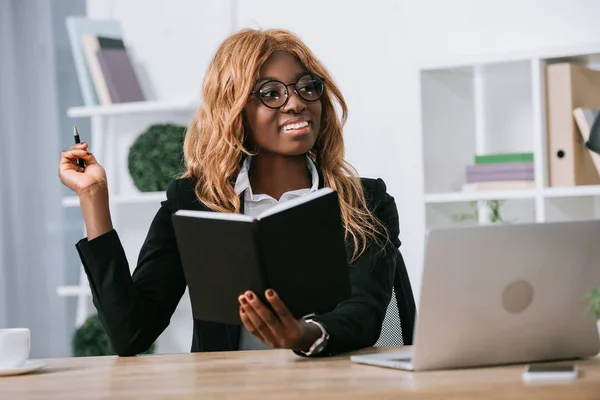 Feliz mujer de negocios afroamericana sentada en gafas y sosteniendo el cuaderno y la pluma - foto de stock