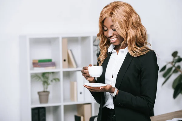 Feliz Africano americano empresária segurando xícara de café no escritório moderno — Fotografia de Stock