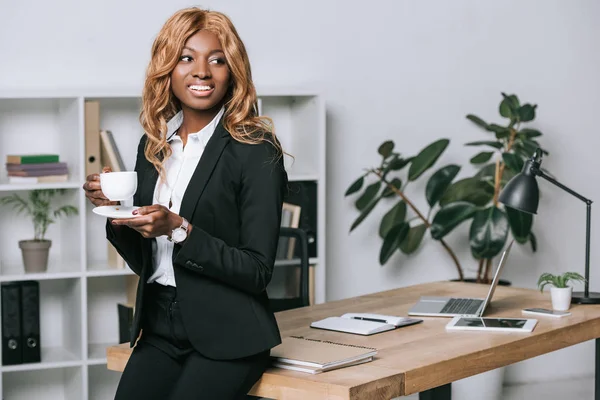 Atractiva mujer de negocios afroamericana sonriendo con una taza de café en la oficina moderna - foto de stock