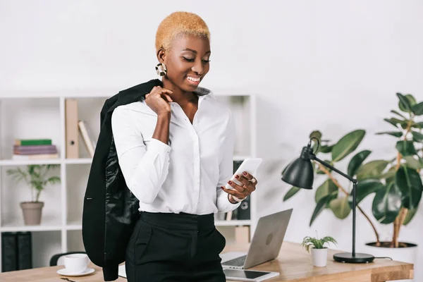 Африканская американская бизнесвумен с короткими волосами, смотрящая на смартфон и держа куртку — стоковое фото