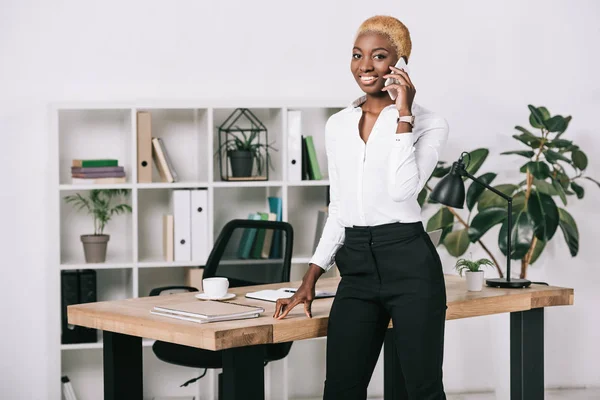 Привлекательная африканская американская бизнесвумен с короткими волосами разговаривающая на смартфоне в современном офисе — стоковое фото
