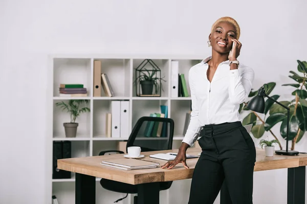 Щаслива афроамериканська бізнес-леді з коротким волоссям розмовляє на смартфоні в сучасному офісі — стокове фото