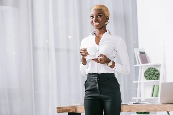 Atractiva mujer de negocios afroamericana con pelo corto sosteniendo taza con bebida en la oficina moderna - foto de stock