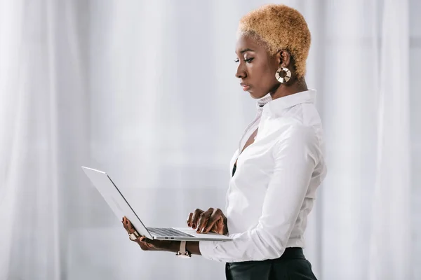 Donna d'affari afroamericana fiduciosa con capelli corti in piedi e utilizzando il computer portatile in ufficio moderno — Foto stock