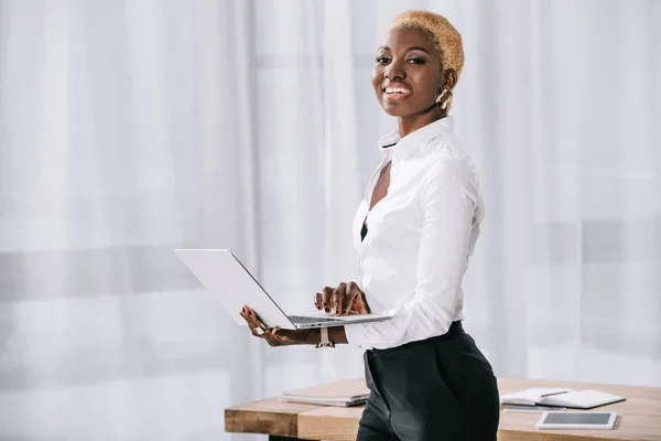 Mujer de negocios afroamericana feliz con el pelo corto de pie y el uso de la computadora portátil en la oficina moderna - foto de stock