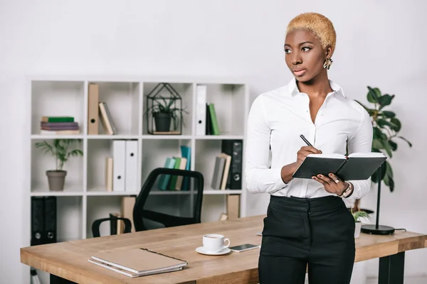 Pensativa mulher de negócios afro-americana com cabelo curto em pé perto da mesa e escrevendo em notebook — Fotografia de Stock