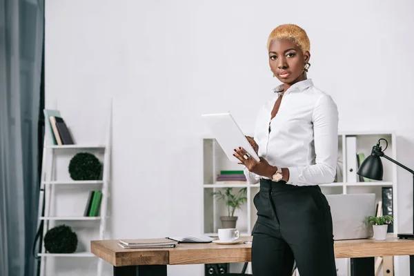 Selbstbewusste afrikanisch-amerikanische Geschäftsfrau mit kurzen Haaren steht am Tisch mit digitalem Tablet — Stockfoto