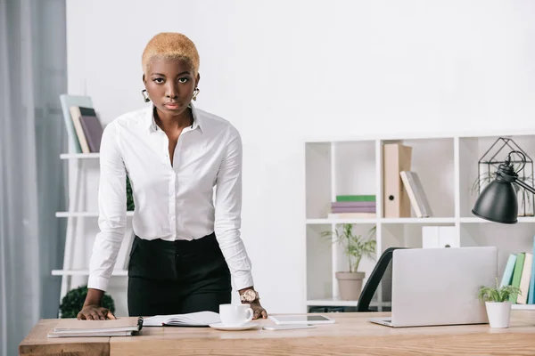 Confiante Africano americano empresária com cabelo curto em pé perto da mesa no escritório moderno — Stock Photo
