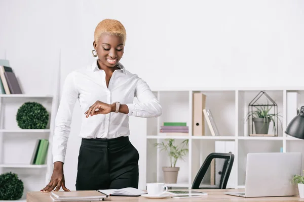 Africano americano empresária com cabelo curto olhando para assistir no escritório moderno — Fotografia de Stock