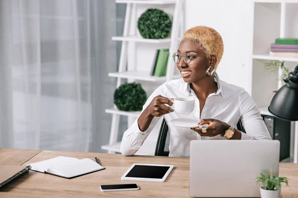 Mujer de negocios afroamericana con pelo corto sosteniendo taza con bebida en la oficina moderna - foto de stock