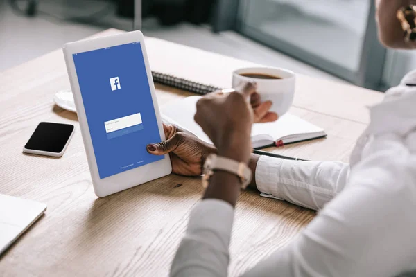 Cropped view of african american woman holding digital tablet with facebook app and cup of coffee — Stock Photo