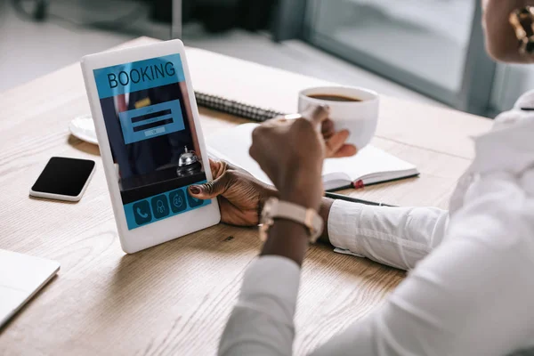 Vista cortada de mulher africana americana segurando copo e tablet digital com aplicativo de reserva na tela — Fotografia de Stock