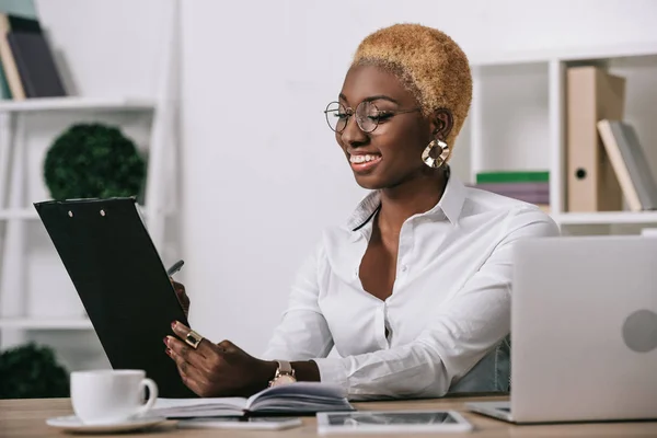 Fröhliche afrikanisch-amerikanische Geschäftsfrau hält Klemmbrett in modernem Büro — Stockfoto