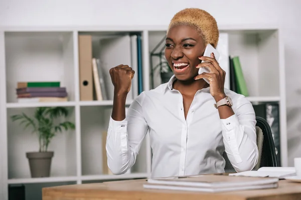 Feliz mujer de negocios afroamericana hablando en el teléfono inteligente y mostrando signo de victoria - foto de stock