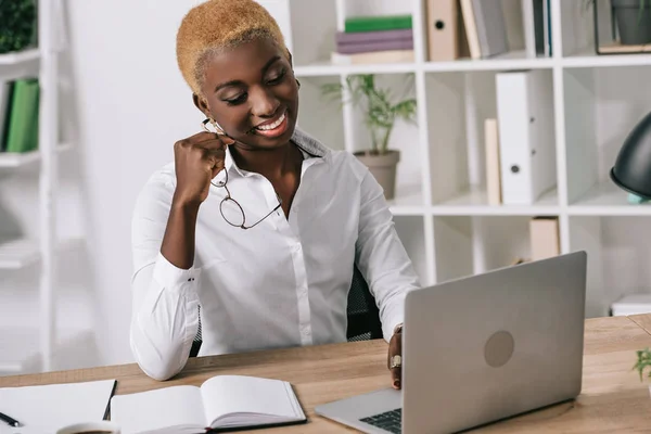 Afrikanisch-amerikanische Geschäftsfrau lächelt und hält Brille neben Laptop — Stockfoto