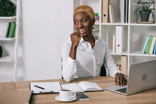 Joyeuse femme d'affaires afro-américaine souriante et tenant des lunettes près d'un ordinateur portable — Photo de stock