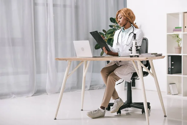 Selbstbewusster afrikanisch-amerikanischer Wissenschaftler sitzt mit Stethoskop und hält Klemmbrett — Stockfoto
