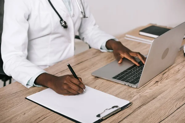 Cropped view of african american scientist writing on paper and typing on laptop — Stock Photo