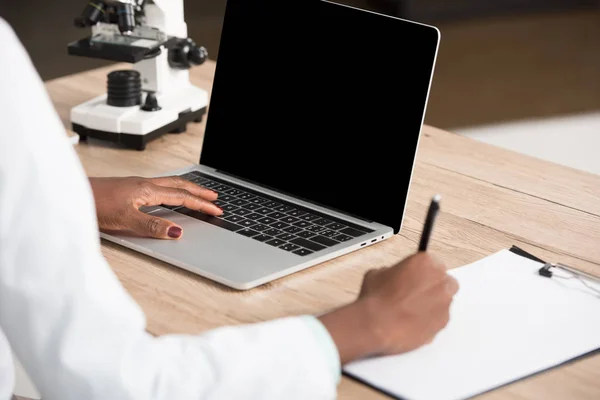 Vista recortada del médico afroamericano escribiendo en papel y usando computadora portátil con pantalla en blanco - foto de stock