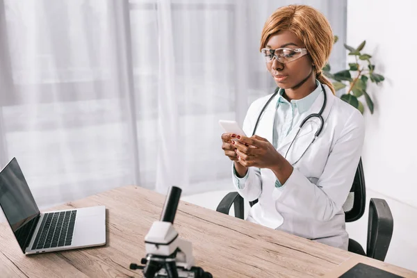 Cientista americano africano atraente sentado em óculos e usando smartphone em laboratório — Fotografia de Stock