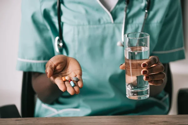 Vista recortada de la enfermera afroamericana con estetoscopio sosteniendo un vaso de agua y pastillas en la mano - foto de stock