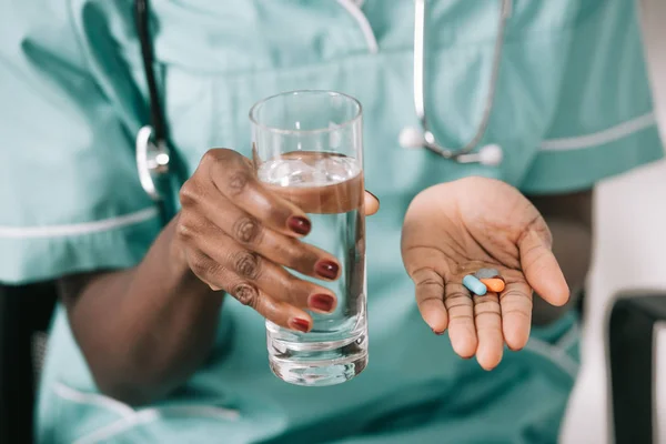 Cropped view of african american nurse with stethoscope holding glass of water and pills in hand — Stock Photo