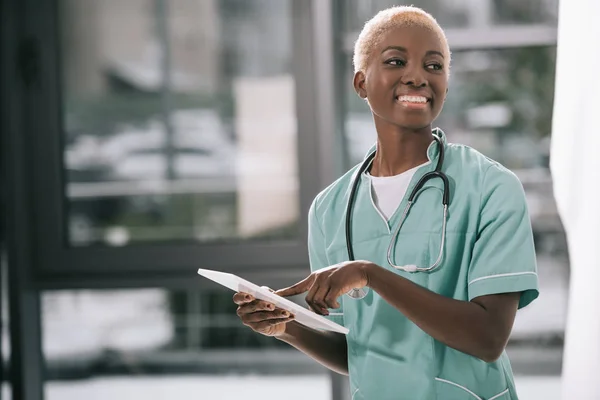 Усміхнена афроамериканська жінка зі стетоскопом за допомогою цифрового планшета в клініці — стокове фото