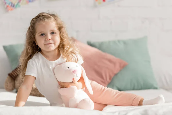 Adorable kid sitting on bed with rabbit toy in children room and looking at camera — Stock Photo